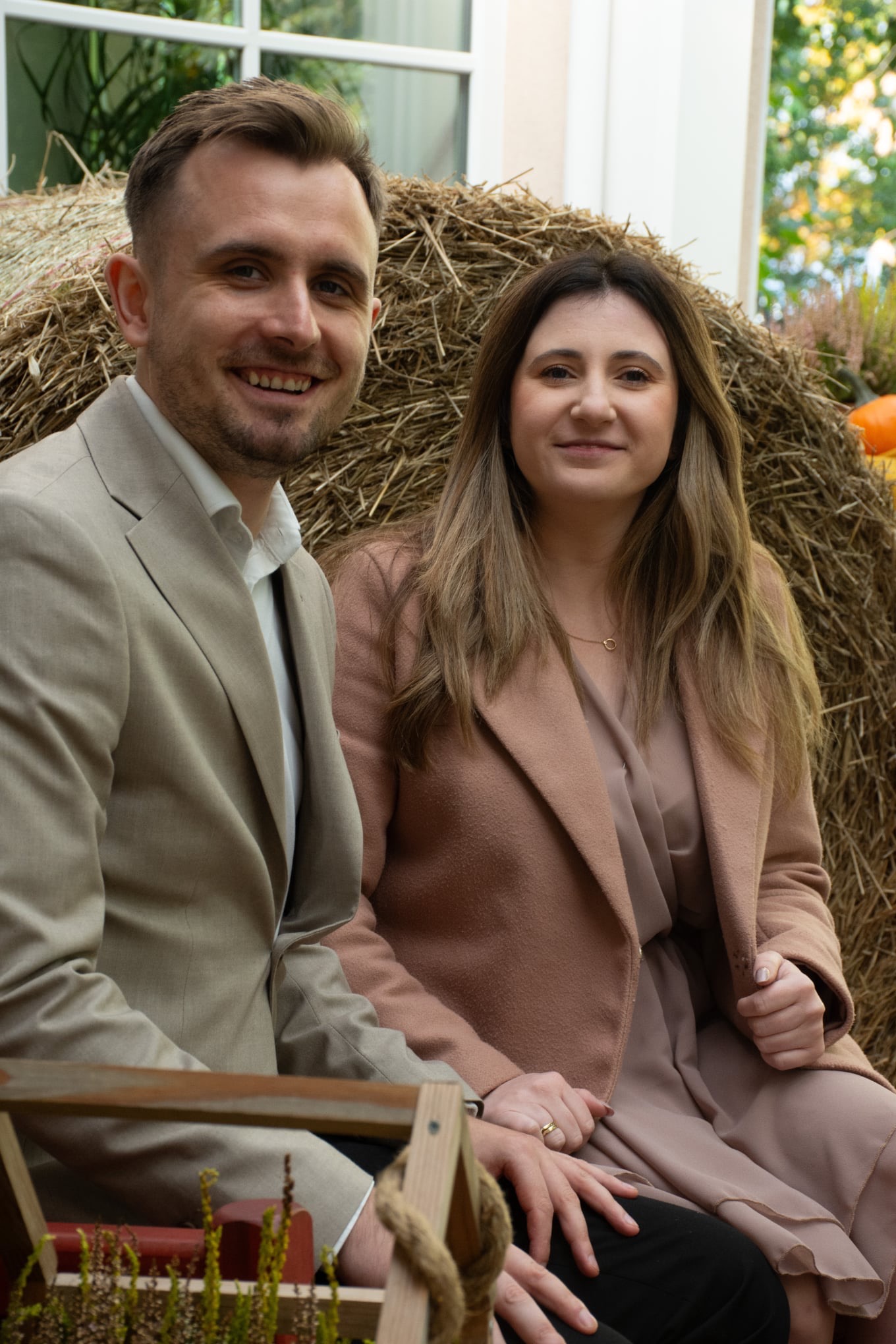Uśmiechnięta para w jesiennej scenerii: mężczyzna w beżowym garniturze i kobieta w płaszczu, siedzą na tle stogu siana z dyniami. Naturalne światło, ciepłe barwy.