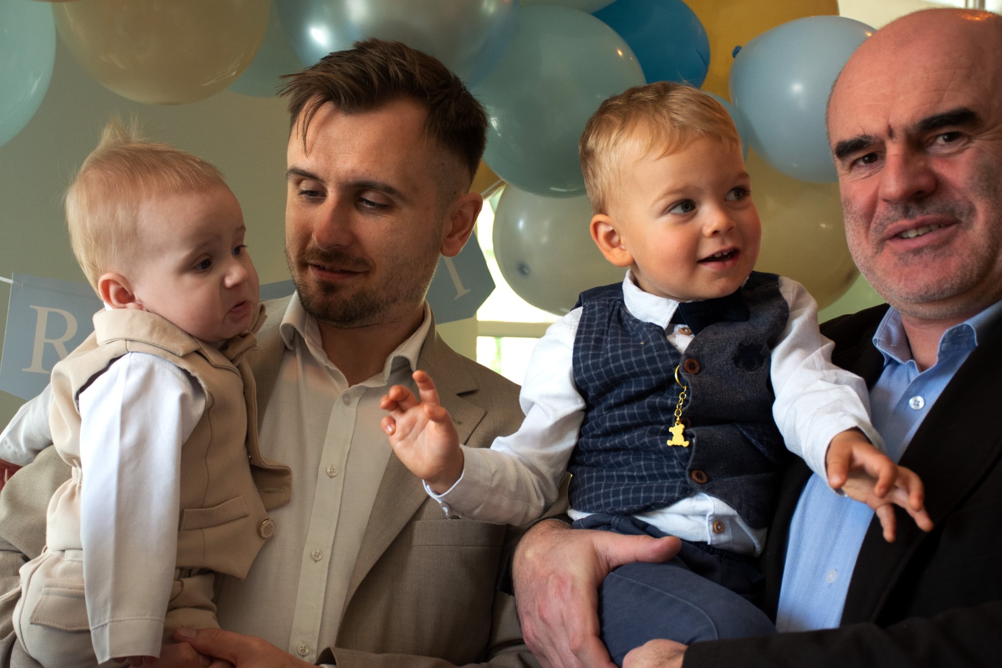 Portret rodzinny: dwóch ojców trzymających synów na tle pastelowych balonów. Jeden z chłopców ma zawieszkę misia. Uśmiechy i eleganckie stroje.