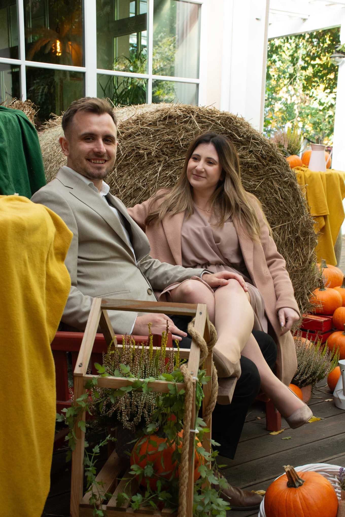 Uśmiechnięta para w jesiennej scenerii: mężczyzna w beżowym garniturze, kobieta w sukience i płaszczu, siedzą na tle siana i dyń. Romantyczna sesja plenerowa.