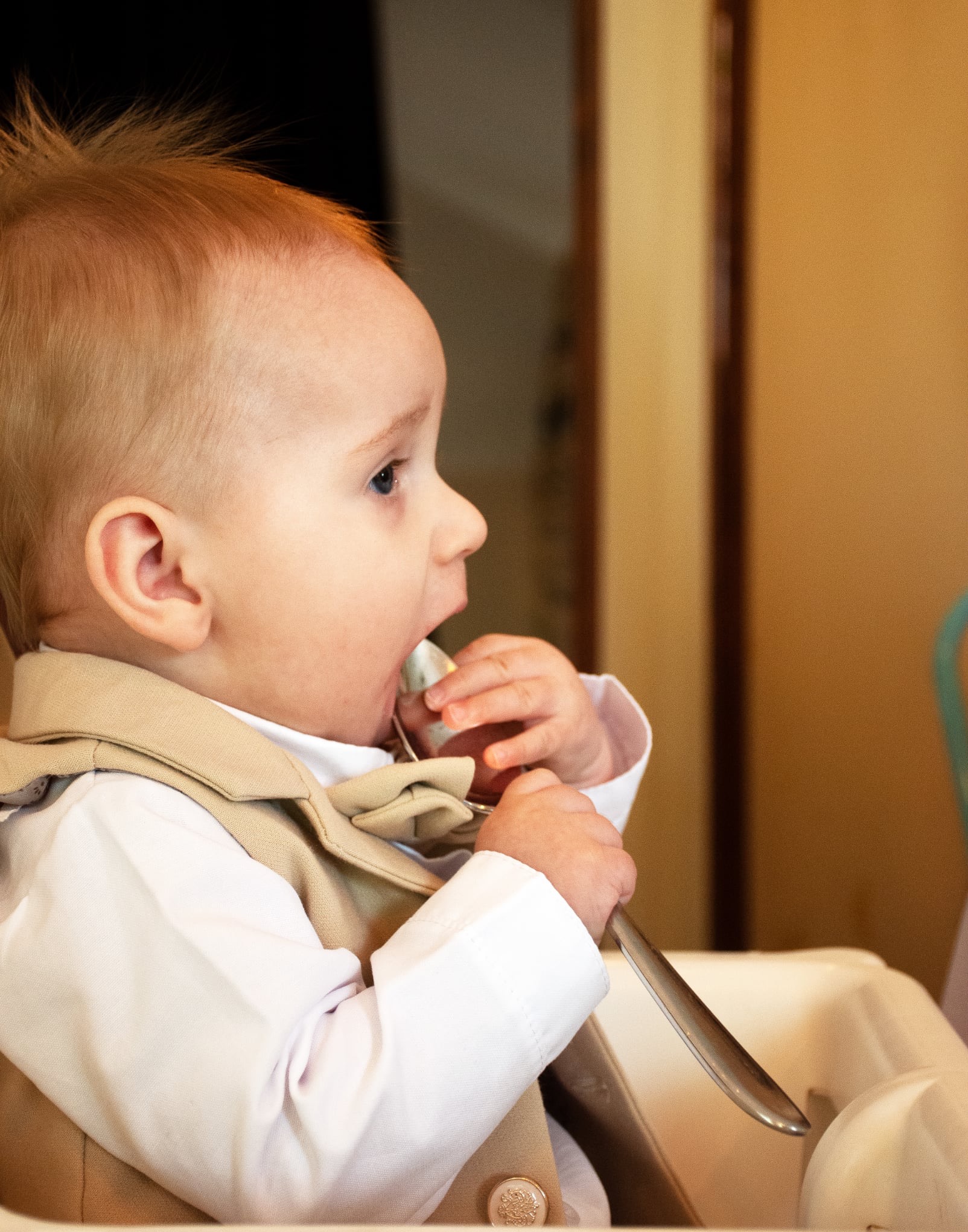 Profil małego chłopca w eleganckiej kamizelce, trzymającego łyżkę w ustach. Jasne włosy i niebieskie oczy dziecka kontrastują z ciepłym tłem.