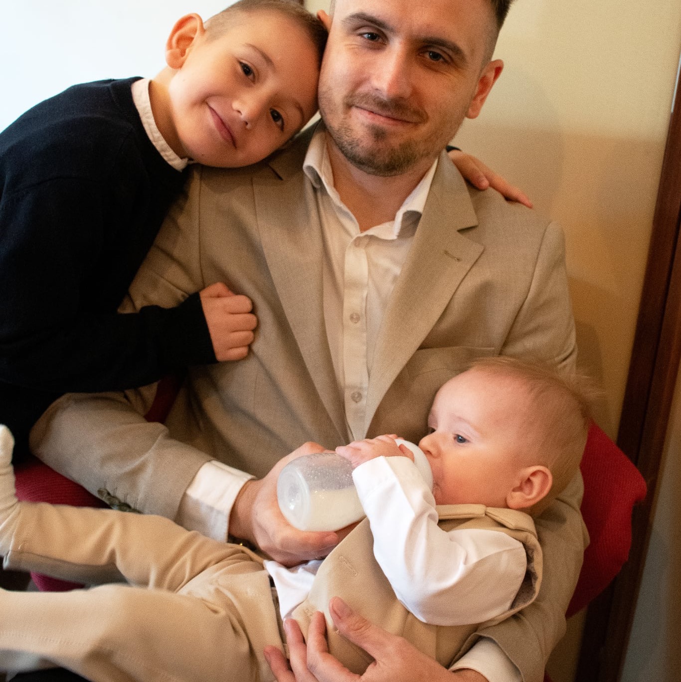 Uśmiechnięty tata w beżowym garniturze karmi butelką niemowlę, a starszy syn opiera się o jego ramię. Ciepłe, naturalne światło.