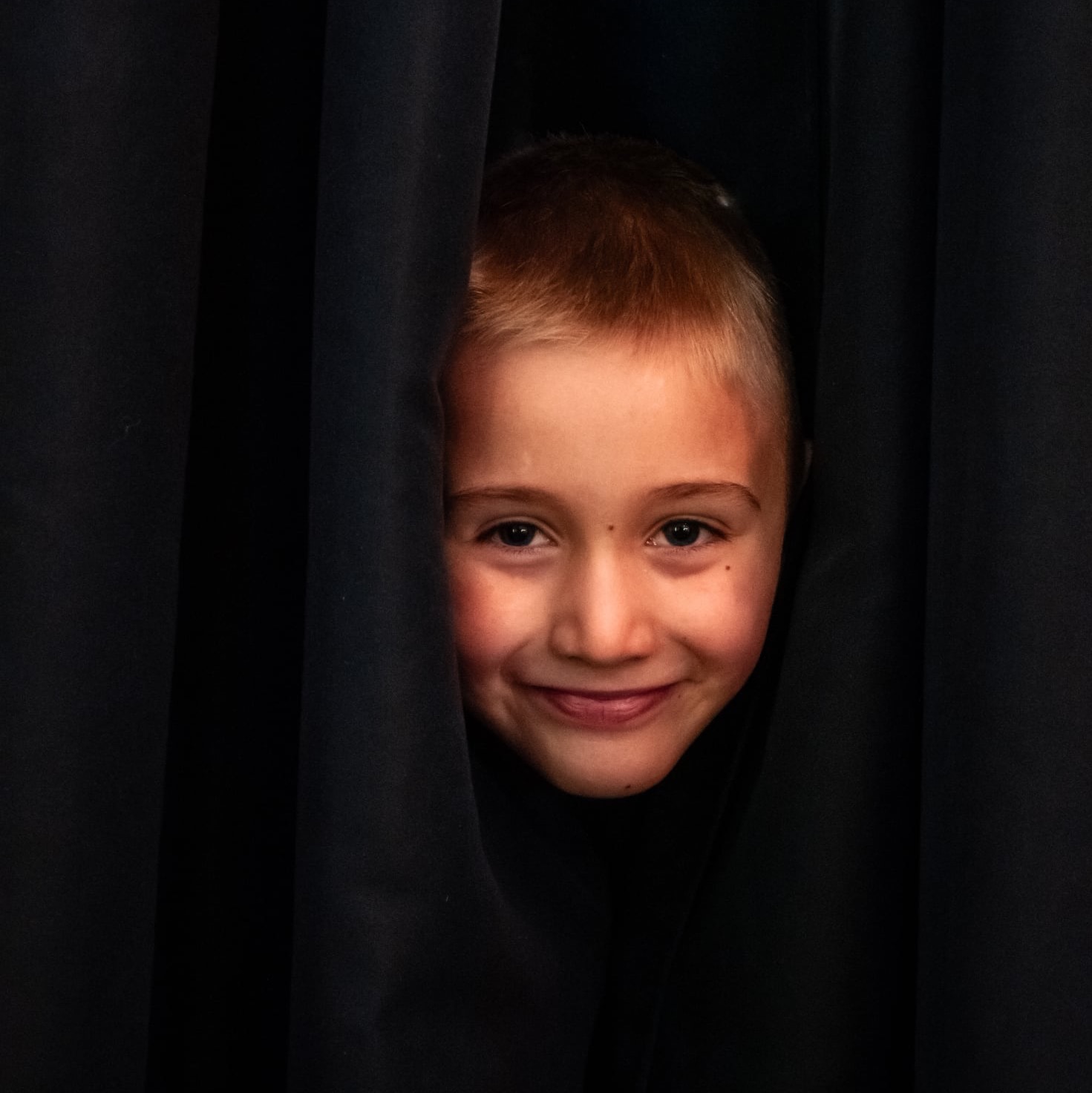 Uśmiechnięty chłopiec z piegami, wyłaniający się zza czarnych zasłon, w kadrze portretowym z bliska, z delikatnym oświetleniem.