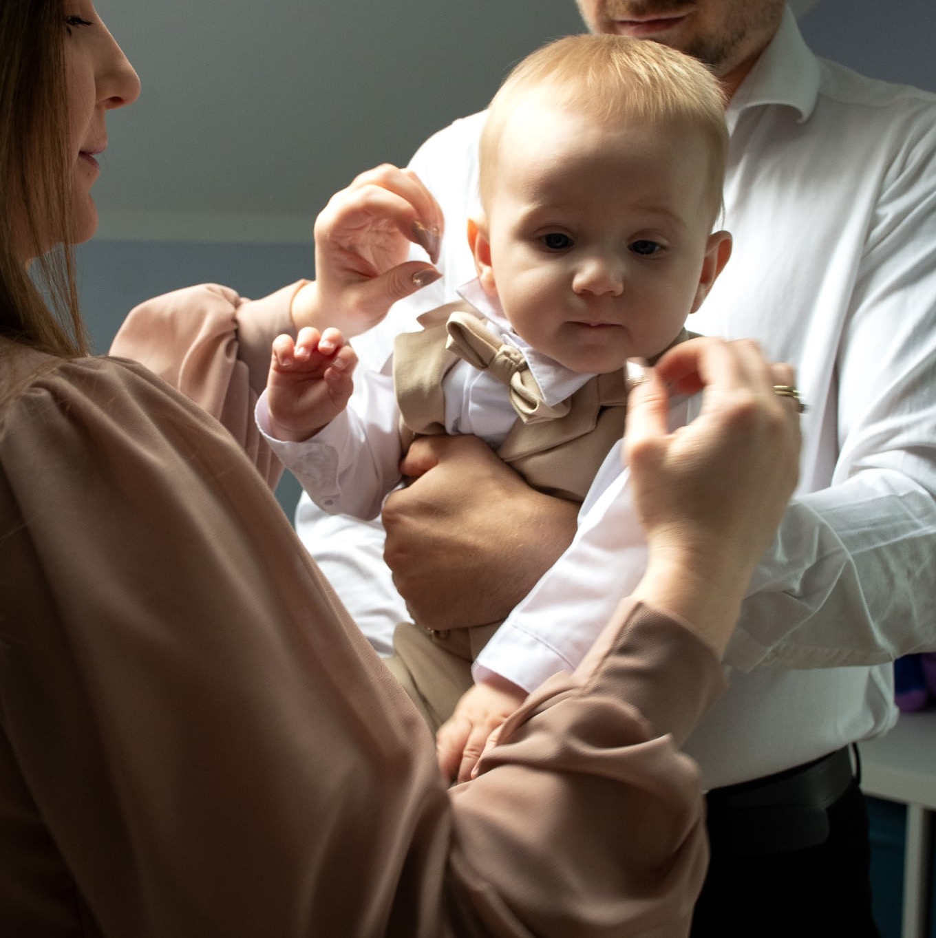 Rodzice ubierają małego chłopca w beżowy garnitur z muszką. Przygotowania do uroczystości rodzinnej, delikatne światło, bliski kadr.
