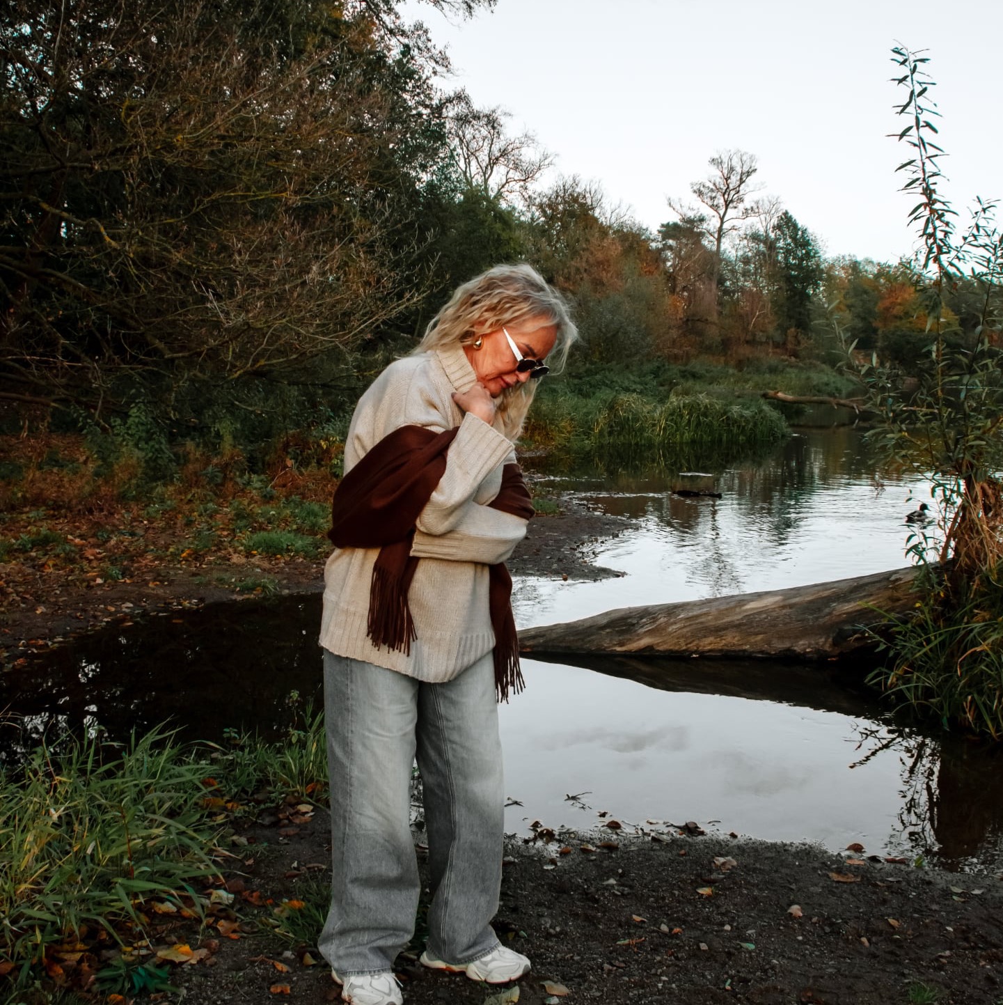 Stylowa kobieta w okularach, beżowym swetrze i szarych jeansach pozuje na tle jesiennego krajobrazu z jeziorem. Uwagę przykuwa brązowy szal z frędzlami.