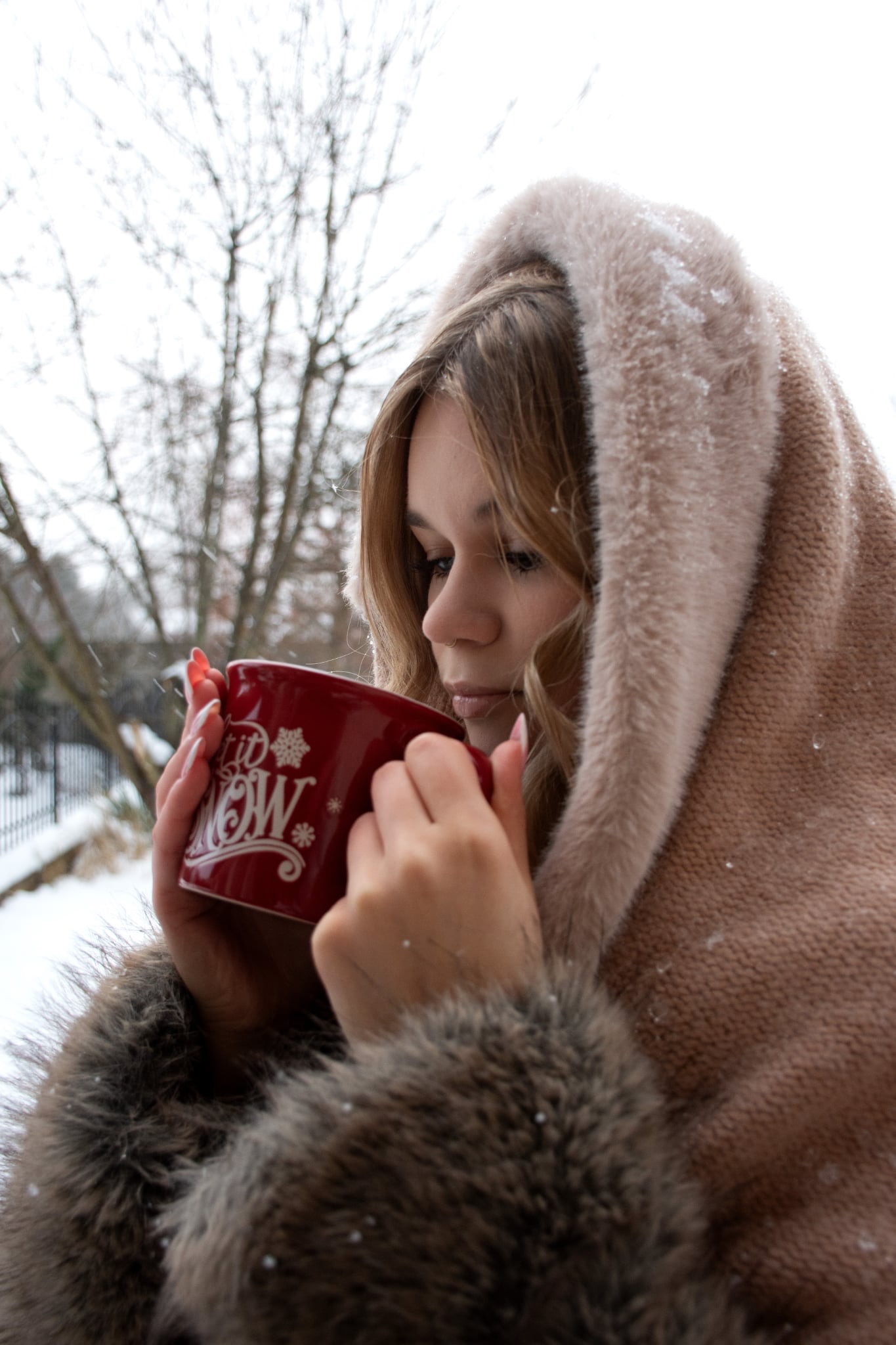 Zimowa sesja portretowa: blondynka w futrzanym okryciu z kapturem pije z czerwonego kubka na tle zaśnieżonych drzew. Delikatne światło podkreśla profil twarzy.
