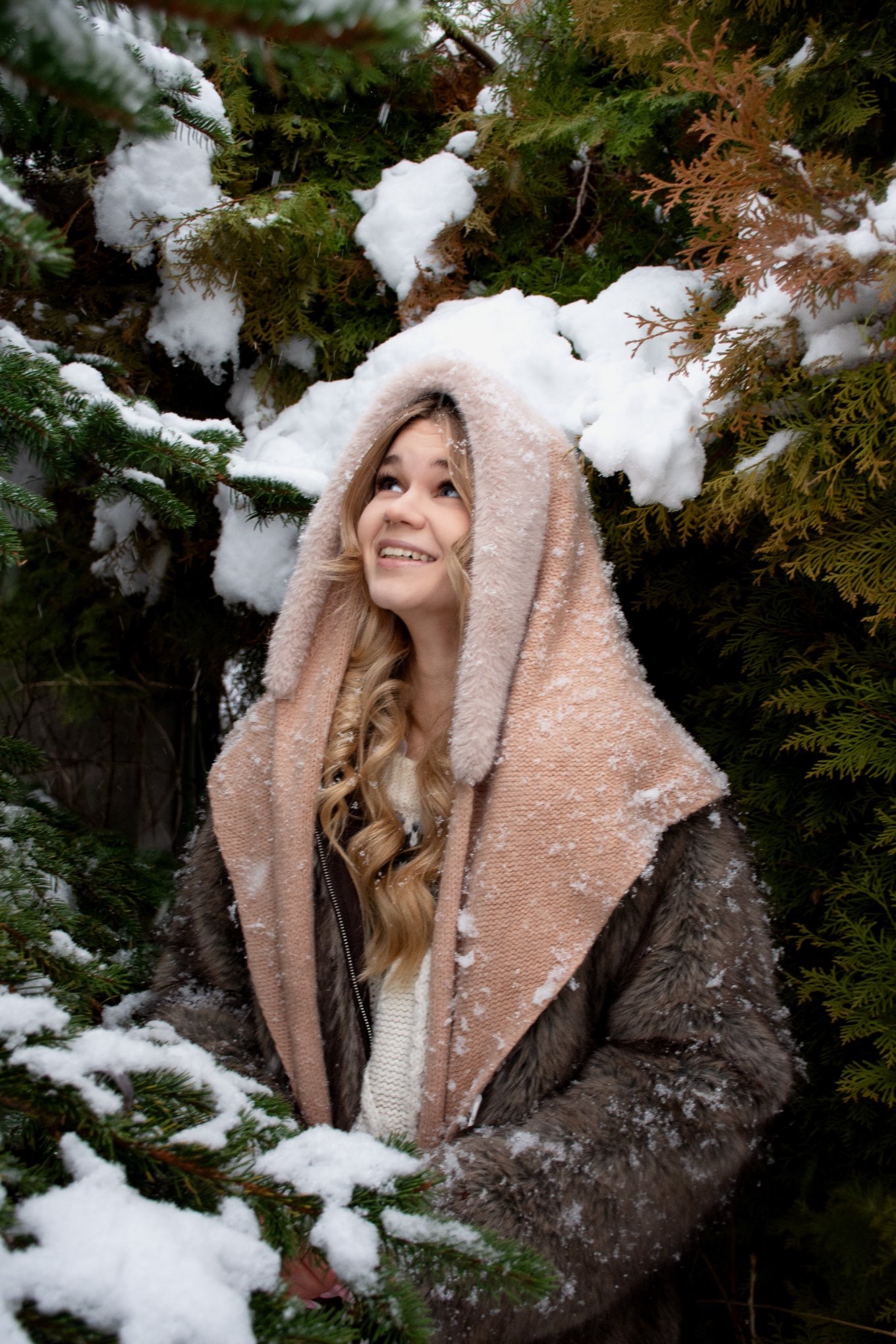 Uśmiechnięta blondynka w futrze i kapturze, otoczona ośnieżonymi gałązkami drzew iglastych, spogląda w górę w zimowej scenerii. Portret w plenerze.