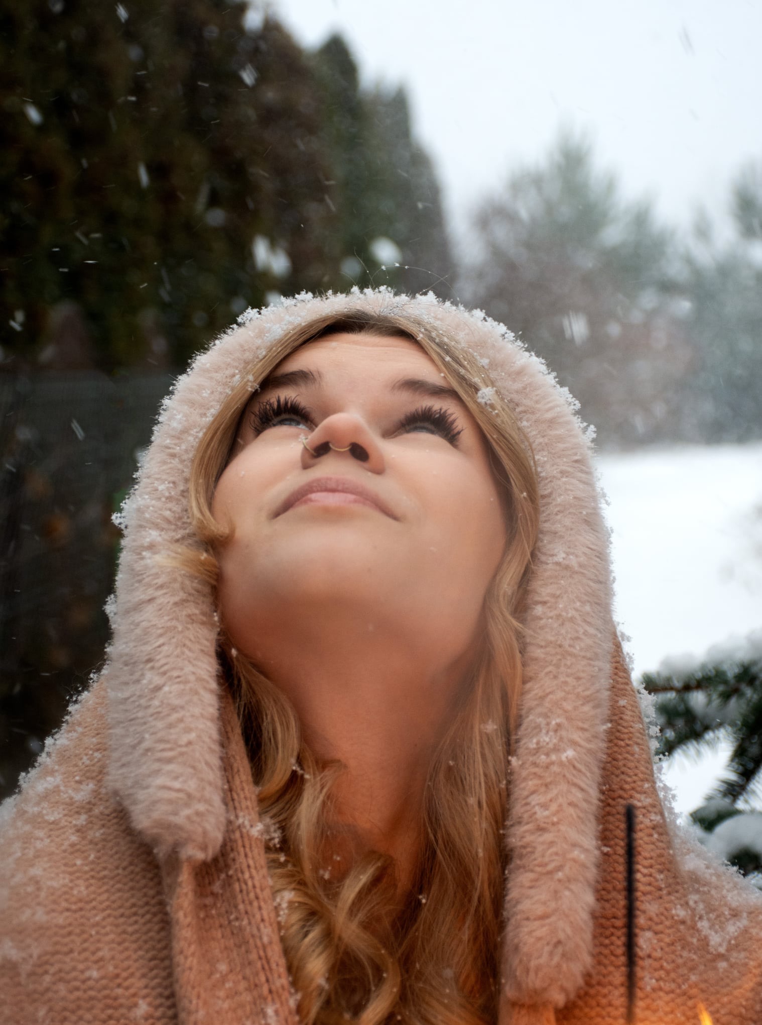 Młoda kobieta z nosem ozdobionym kolczykiem, w kapturze obsypanym śniegiem, spogląda w górę na padający śnieg. Ujęcie z dołu, zimowa sceneria w tle.