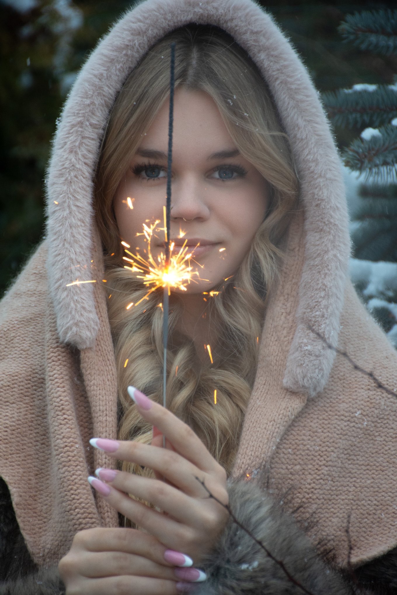 Młoda kobieta z blond włosami w kapturze trzyma płonącą zimną iskrę na tle zimowego krajobrazu. Widoczne płatki śniegu na kapturze i gałęziach drzew.