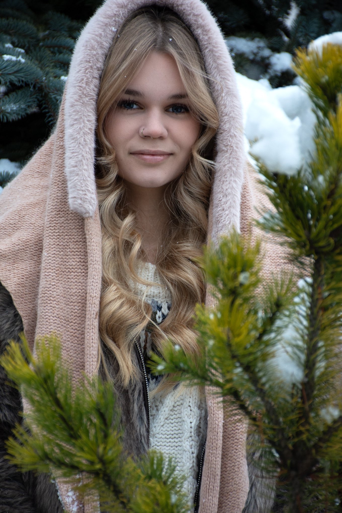 Portret młodej kobiety z kolczykiem w nosie, w zimowej scenerii, ubrana w ciepły szal z kapturem, z delikatnym uśmiechem, na tle ośnieżonych gałęzi drzew iglastych.