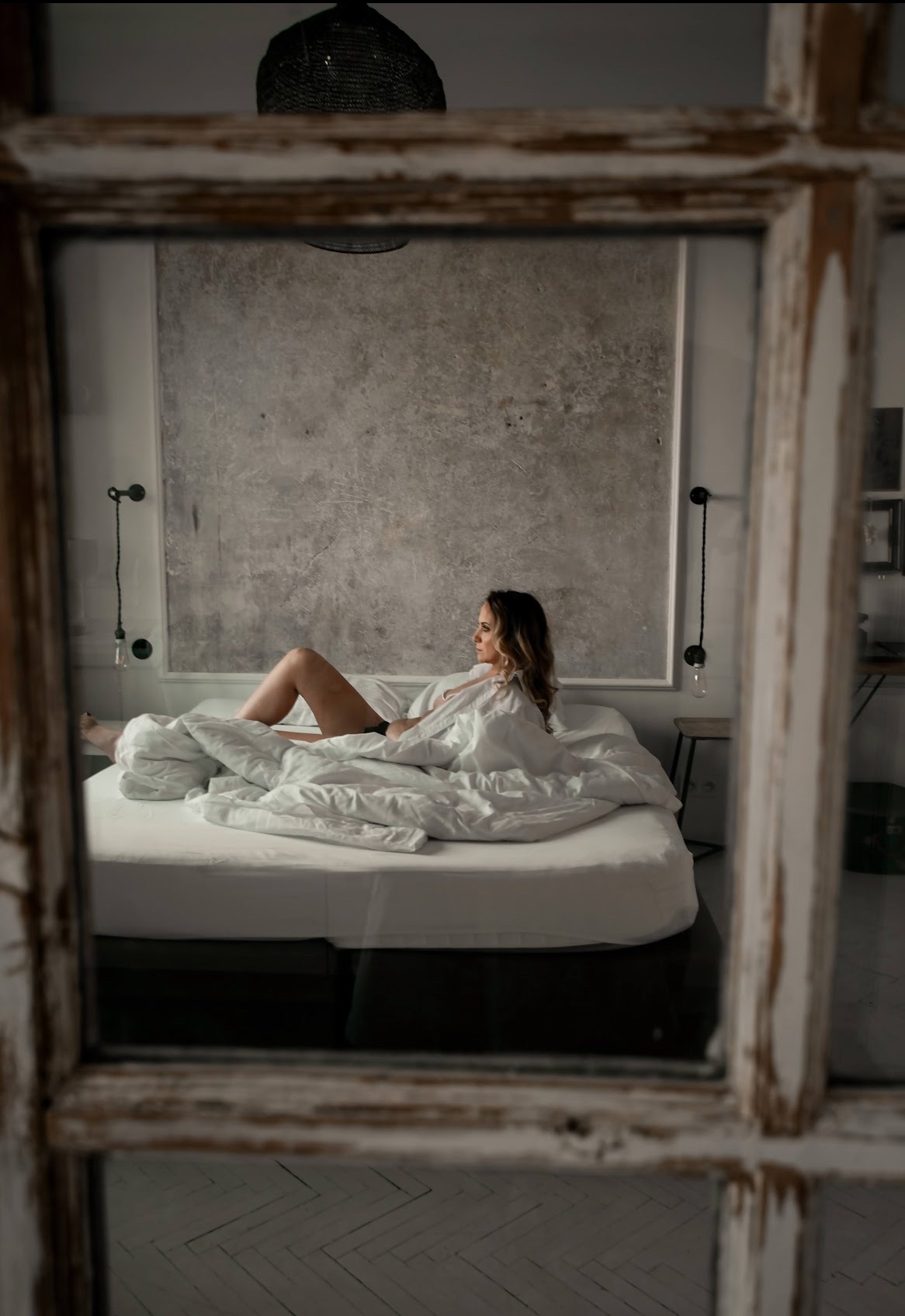 Kobieta w białej koszuli leży na łóżku, widziana przez starą, drewnianą ramę okienną. Stonowana kolorystyka, naturalne światło, minimalistyczny wystrój sypialni.