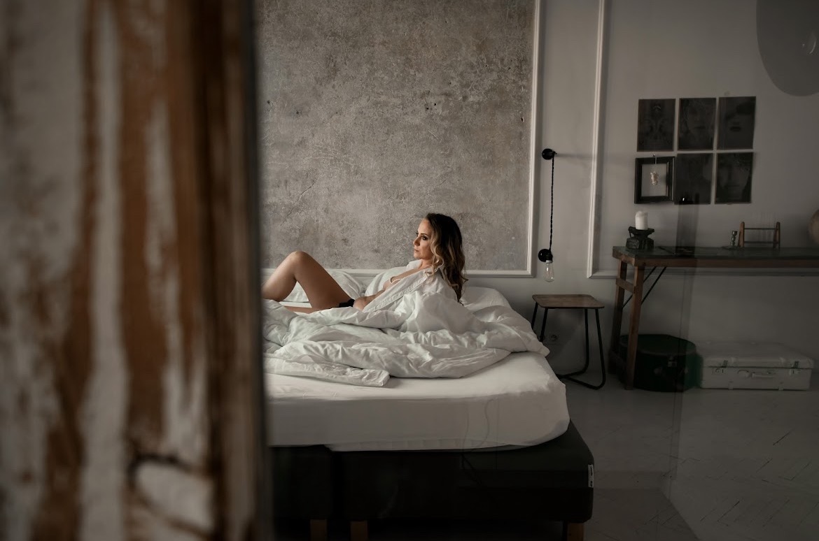 Kobieta w białej koszuli leży na łóżku w pokoju z betonową ścianą. Ujęcie zza drewnianej framugi, z boku, w stonowanych barwach.