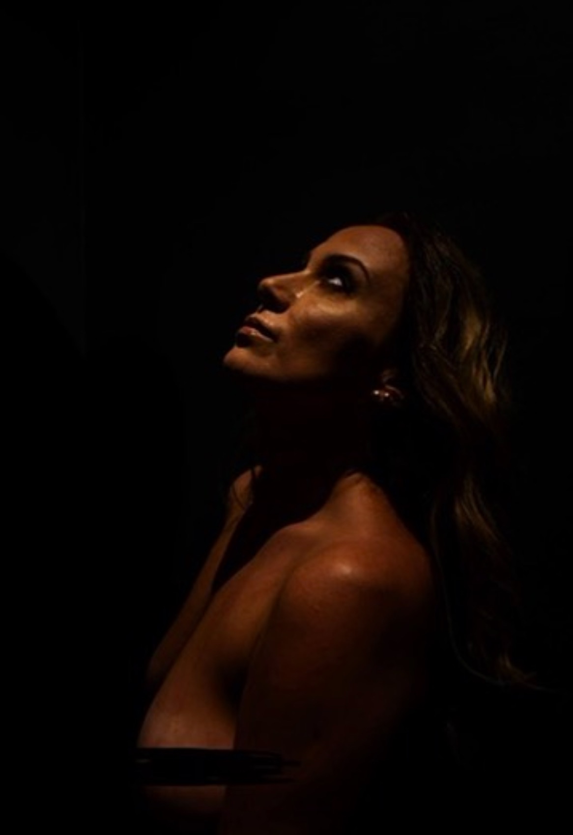 Kobiecy akt w kontraście światła i cienia, z profilu, z głową uniesioną ku górze. Subtelne oświetlenie podkreśla rysy twarzy i linię ramion. Mroczny, intymny klimat.