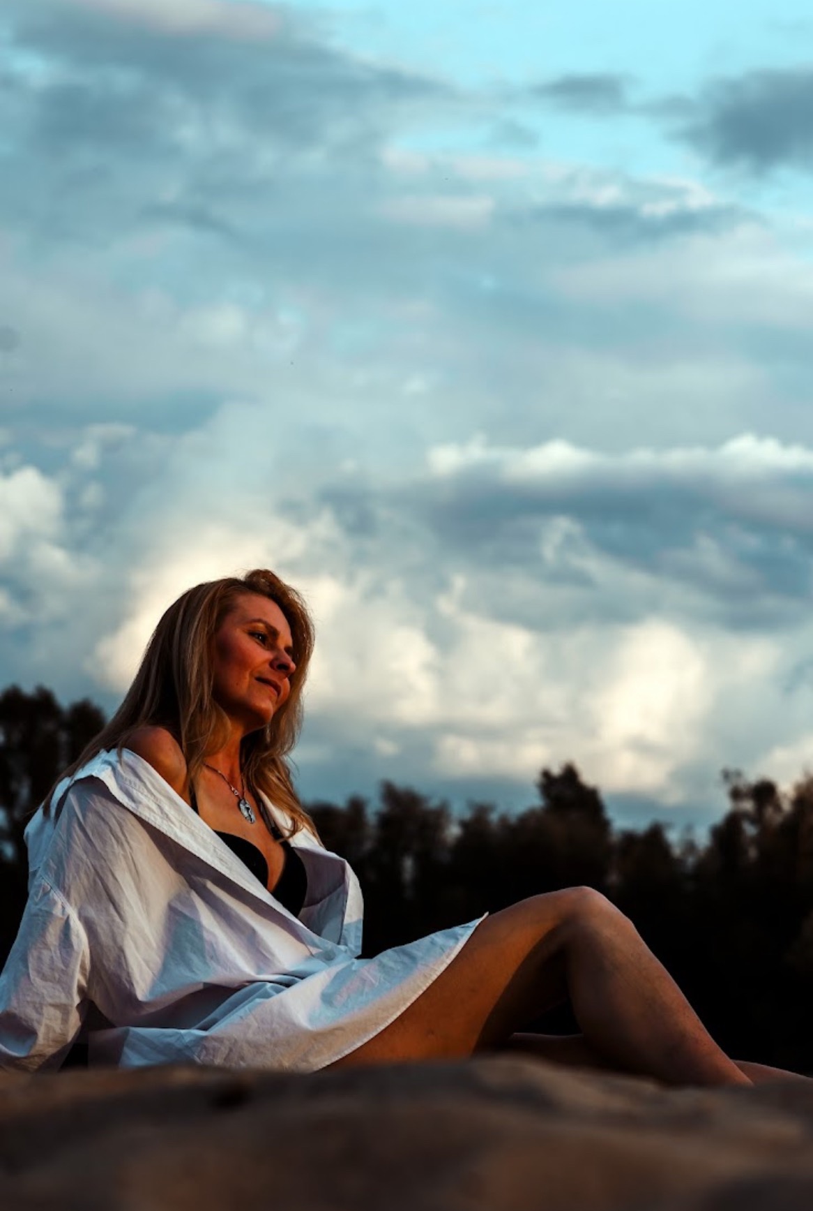 Kobieta w rozpiętej białej koszuli i biustonoszu, siedząca na piasku na tle pochmurnego nieba, w trakcie sesji zdjęciowej.
