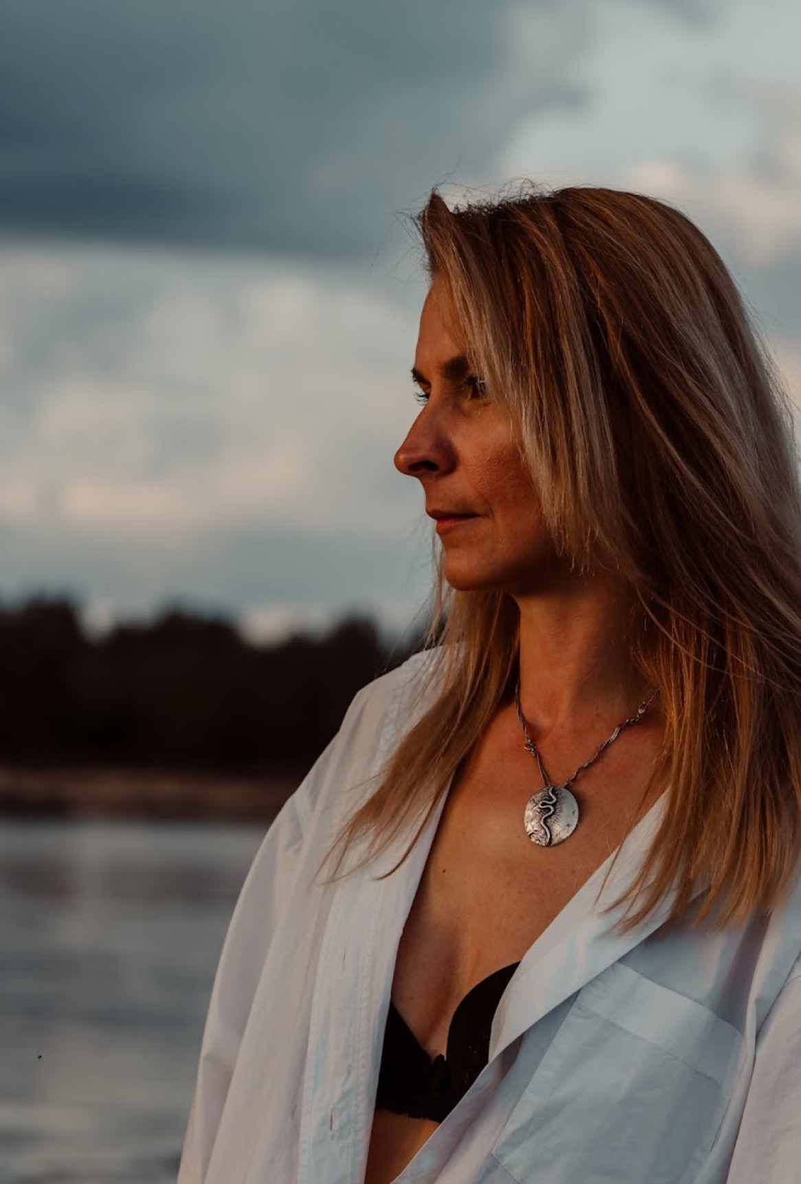 Portret kobiety z profilu, w białej koszuli, z naszyjnikiem na tle zachmurzonego nieba i wody. Delikatne światło podkreśla rysy twarzy i detale biżuterii.