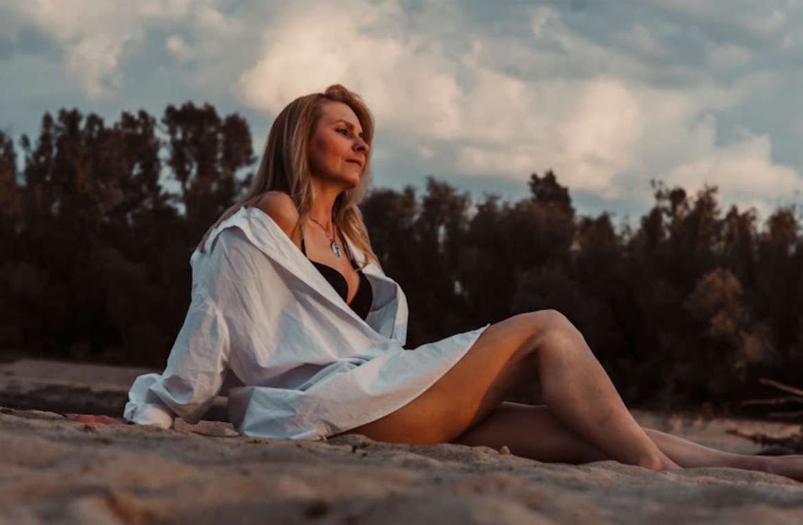 Kobieta w białej koszuli i czarnym bikini siedzi na piaszczystej plaży, zamyślona, z widokiem na zachmurzone niebo i drzewa w tle, w ciepłym, naturalnym oświetleniu.