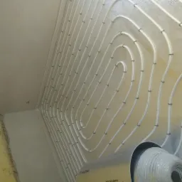 Białe rurki ogrzewania podłogowego ułożone spiralnie na folii termoizolacyjnej w trakcie montażu, widok z dołu, surowy stan wykończenia wnętrza.