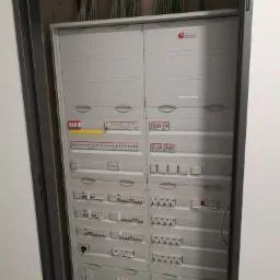 Otwarta szara szafa elektryczna z widocznymi zabezpieczeniami, przełącznikami i przewodami u góry, zamontowana w szarej wnęce ściany.