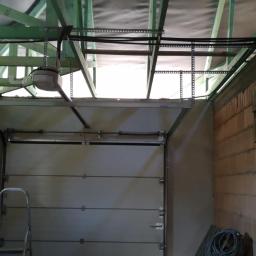 Electric Gods - Montaż automatycznej bramy garażowej w surowym wnętrzu, widoczne przewody elektryczne pod sufitem i drabina oparta o bramę.