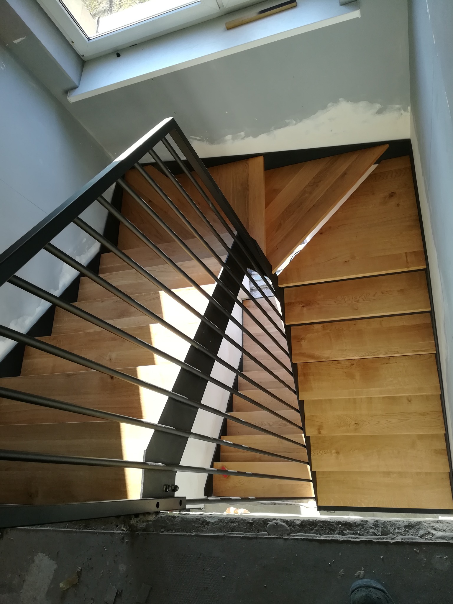 Kątowe ujęcie schodów z drewnianymi stopniami i metalową balustradą w surowym wnętrzu, widoczne ślady prac wykończeniowych.