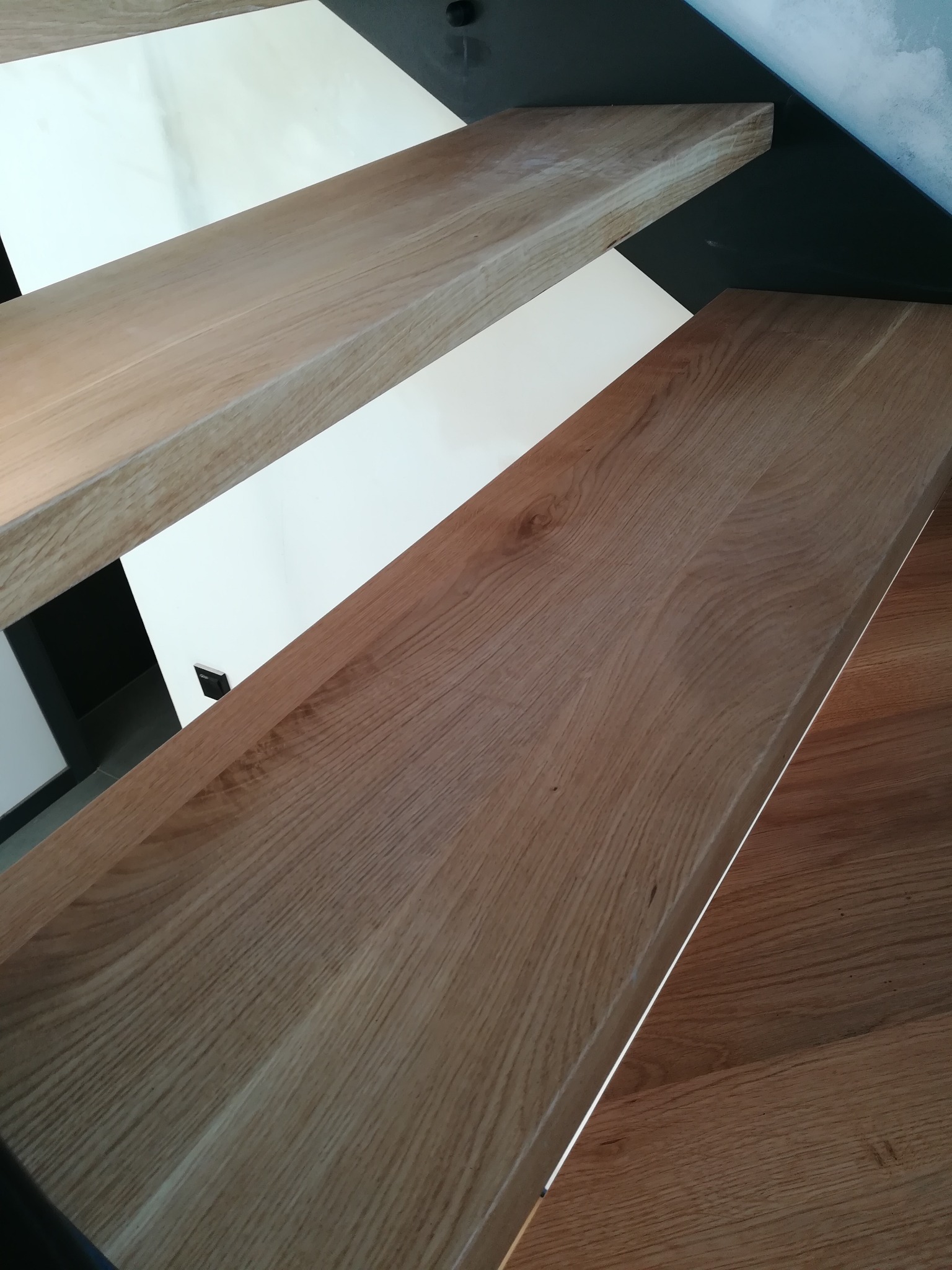 Nowoczesne schody drewniane z jasnego drewna dębowego, z czarnymi metalowymi wspornikami i białymi ścianami, widok z góry na stopnie.