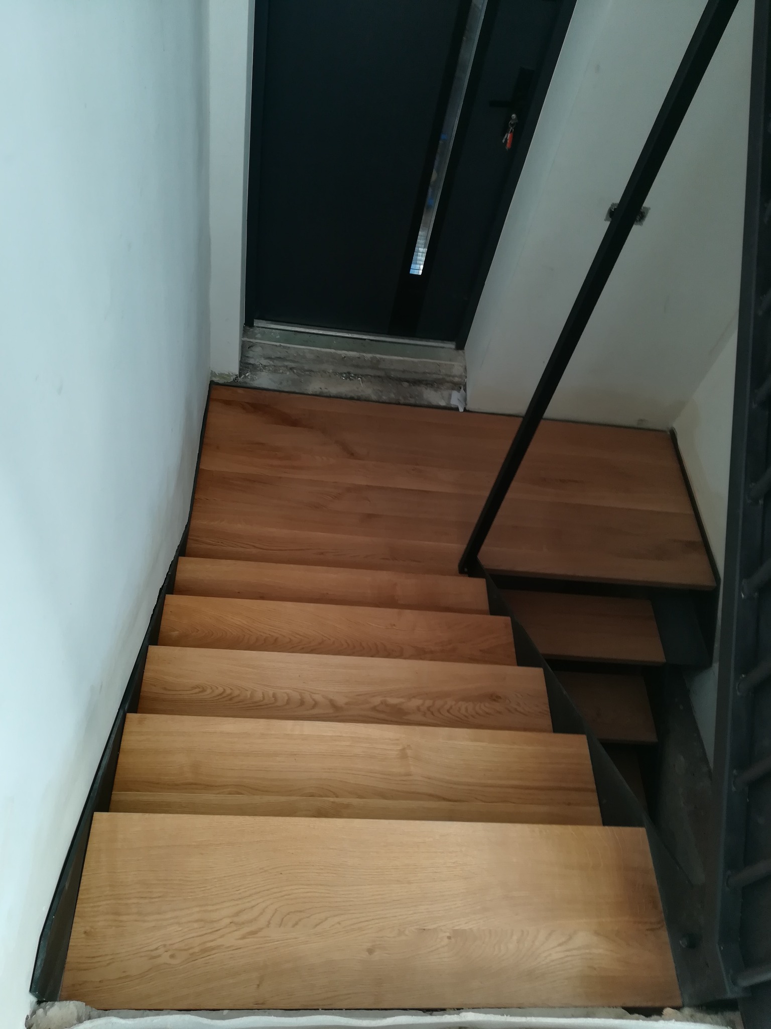 Drewniane schody z metalową balustradą, widok z góry, jasne drewno, czarne elementy metalowe, białe ściany, nowoczesny design.