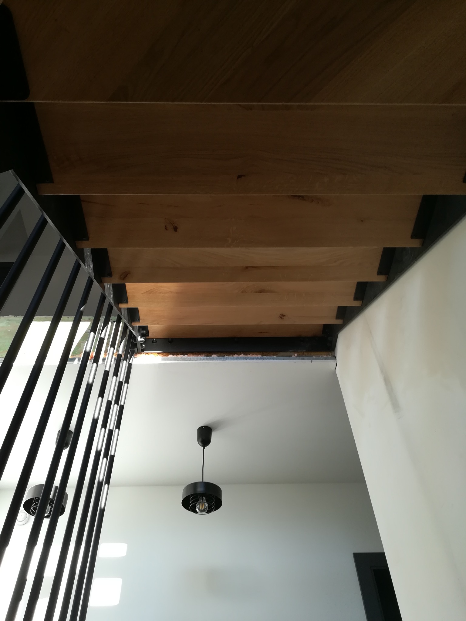 Widok od dołu na drewniane schody z czarnymi metalowymi wspornikami i balustradą, minimalistyczne czarne lampy wiszące na białym suficie, widoczne niedoskonałości wykończenia ścian.