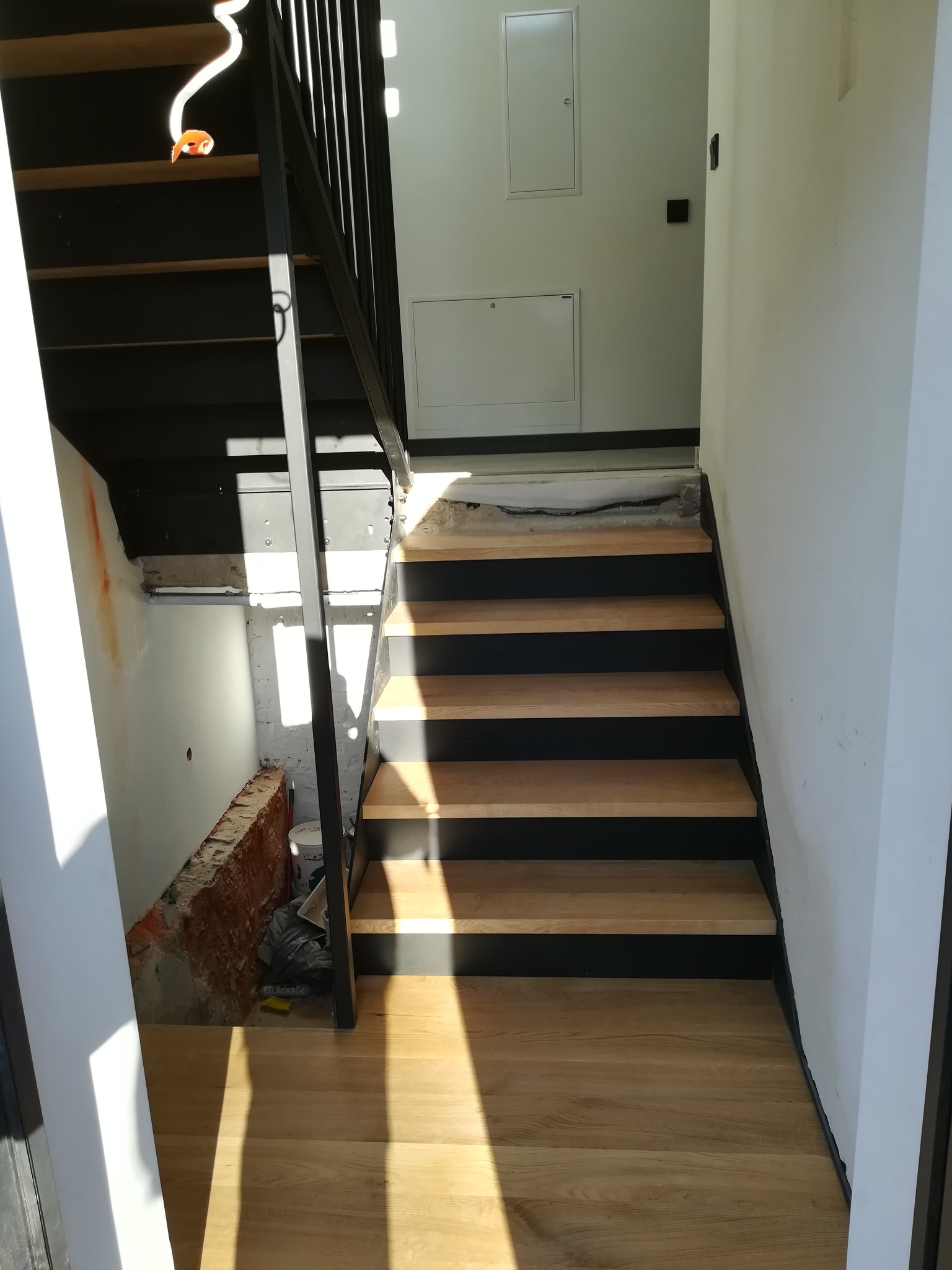 Wewnętrzne schody z drewnianymi stopniami i czarną metalową konstrukcją, widoczne niedokończone prace wykończeniowe przy ścianie.