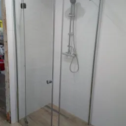 Kabina prysznicowa ze szklanymi drzwiami zamontowana w rogu łazienki, widoczny odpływ liniowy w podłodze wyłożonej płytkami imitującymi drewno, białe płytki na ścianach.