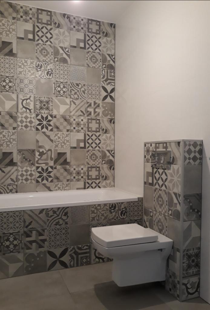 Wykończona łazienka z mozaiką z płytek w odcieniach szarości, obudowa wanny i toaleta podwieszana z obudową również wykończone mozaiką.