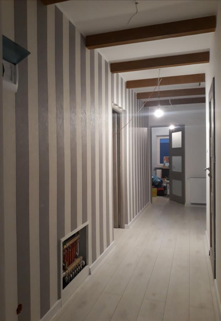 Korytarz po remoncie z tapetą w szaro-białe pionowe pasy, jasną podłogą z paneli i widoczną instalacją grzewczą w ścianie.