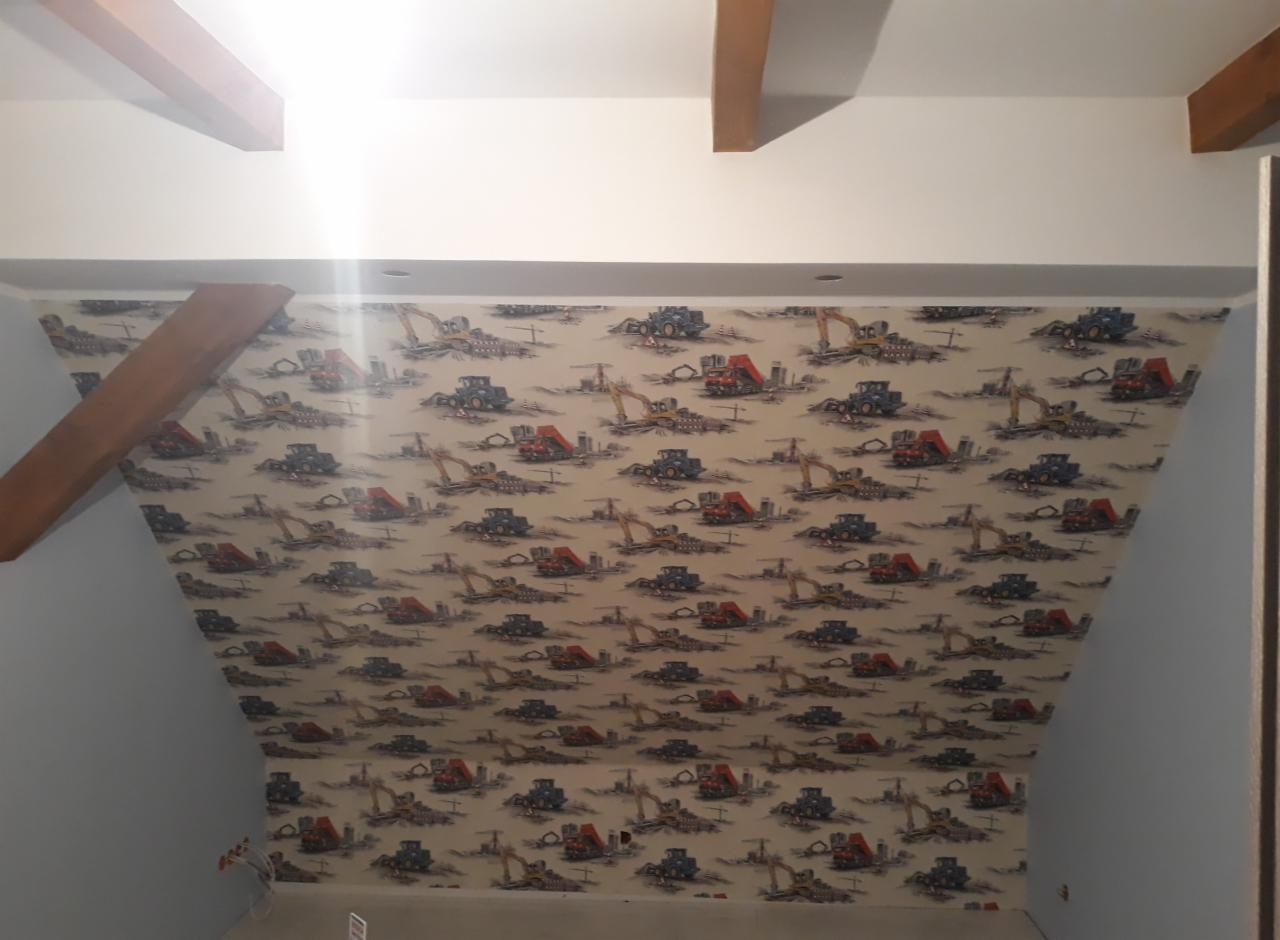 Ściana pod skosem w pokoju, pokryta tapetą z motywem maszyn budowlanych, widoczne elementy drewniane konstrukcji sufitu.