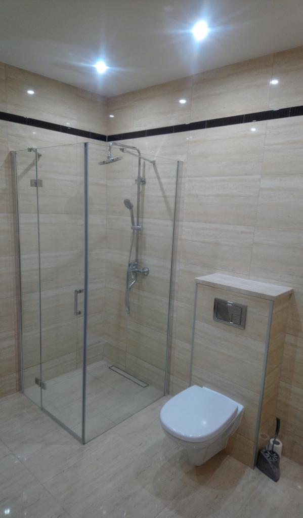 Nowoczesna łazienka z kabiną prysznicową typu walk-in, wiszącą toaletą i beżowymi płytkami imitującymi marmur, oświetlona punktowymi lampami sufitowymi.