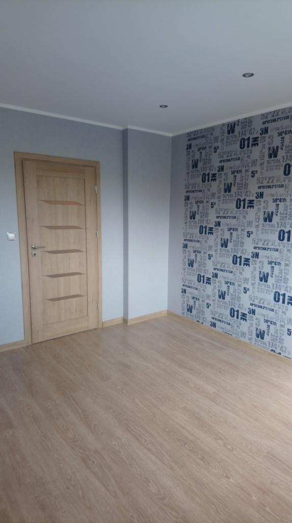 Wykończone wnętrze pokoju z panelami podłogowymi w odcieniach jasnego drewna, drewnianymi drzwiami z poziomymi wgłębieniami i szarą ścianą z wzorem współrzędnych geograficznych.