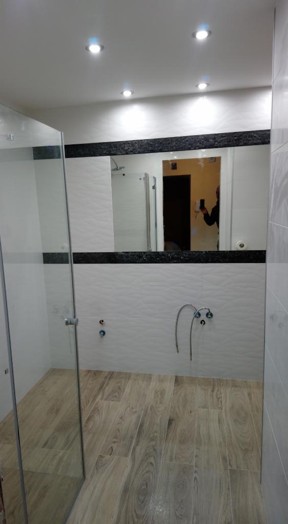 Nowoczesna łazienka z kabiną prysznicową typu walk-in, wykończona płytkami imitującymi drewno na podłodze i białymi płytkami z delikatnym wzorem na ścianach, z czarnym pasem dekoracyjnym pod lustrem.