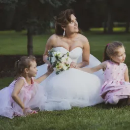 Panna młoda w białej sukni siedzi na trawie z dwiema małymi dziewczynkami w różowych sukienkach, trzyma bukiet kwiatów i patrzy w górę.