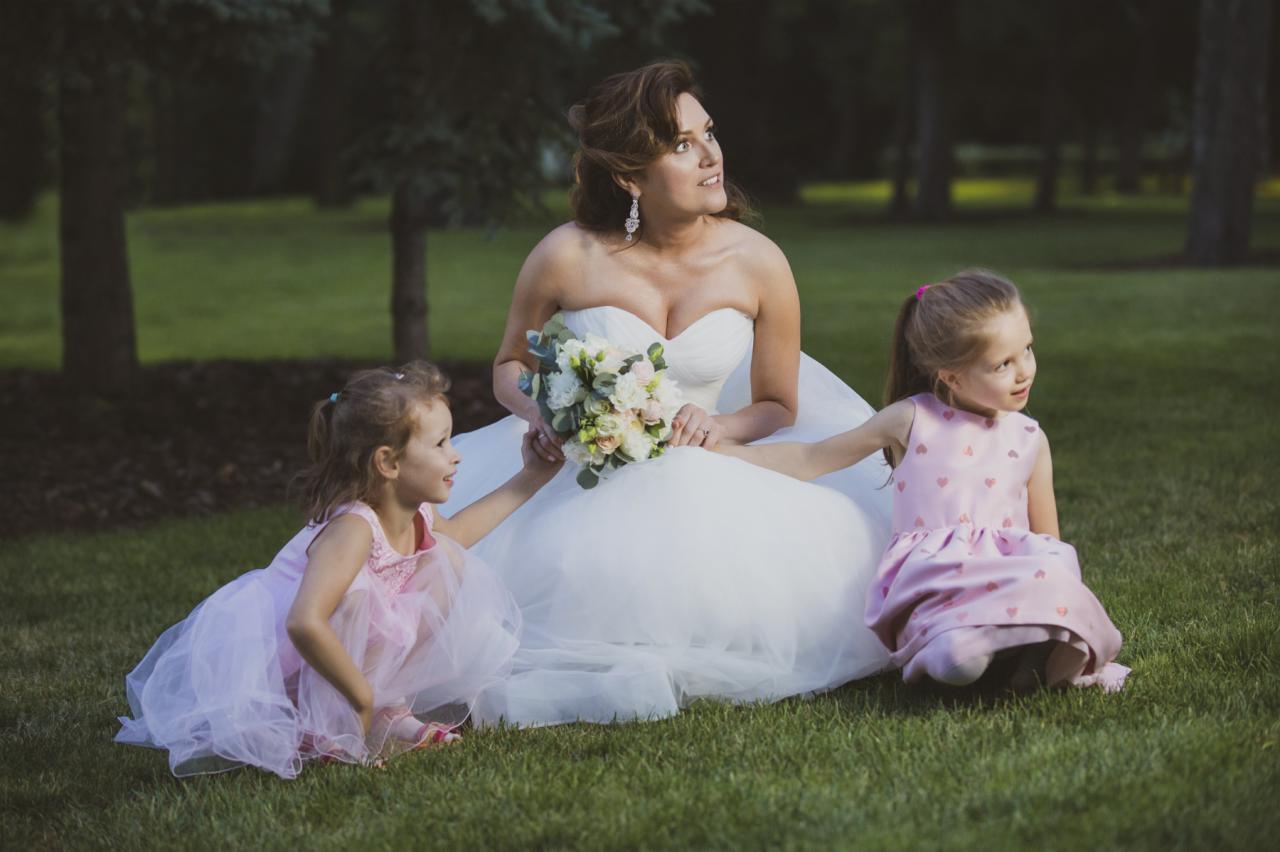 Panna młoda w białej sukni siedzi na trawie z dwiema małymi dziewczynkami w różowych sukienkach, trzyma bukiet kwiatów i patrzy w górę.