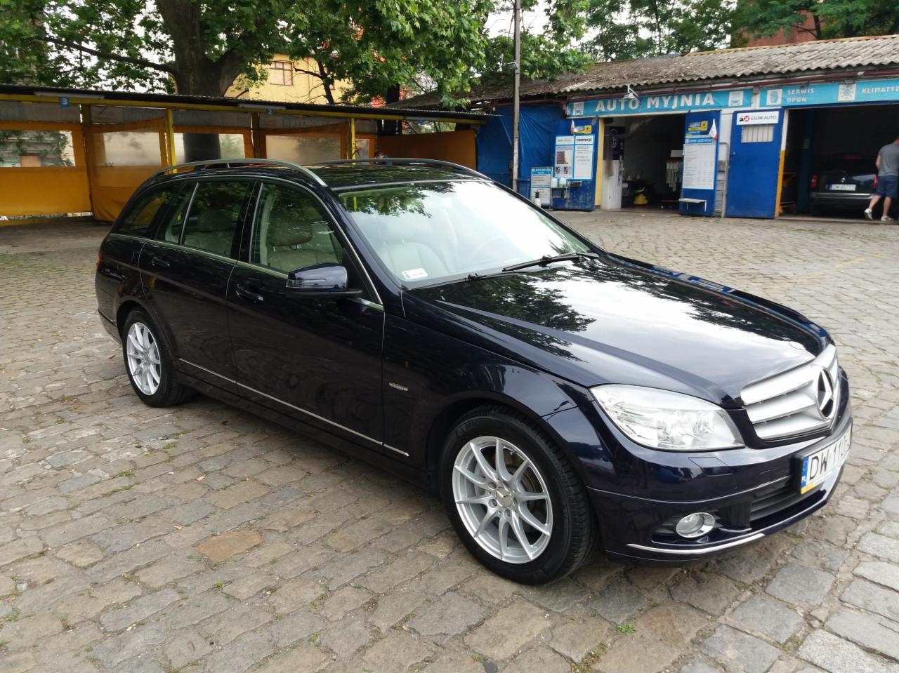 Ciemnogranatowe kombi marki Mercedes stoi na brukowanym placu przed budynkiem z napisem 'AUTO MYJNIA'.