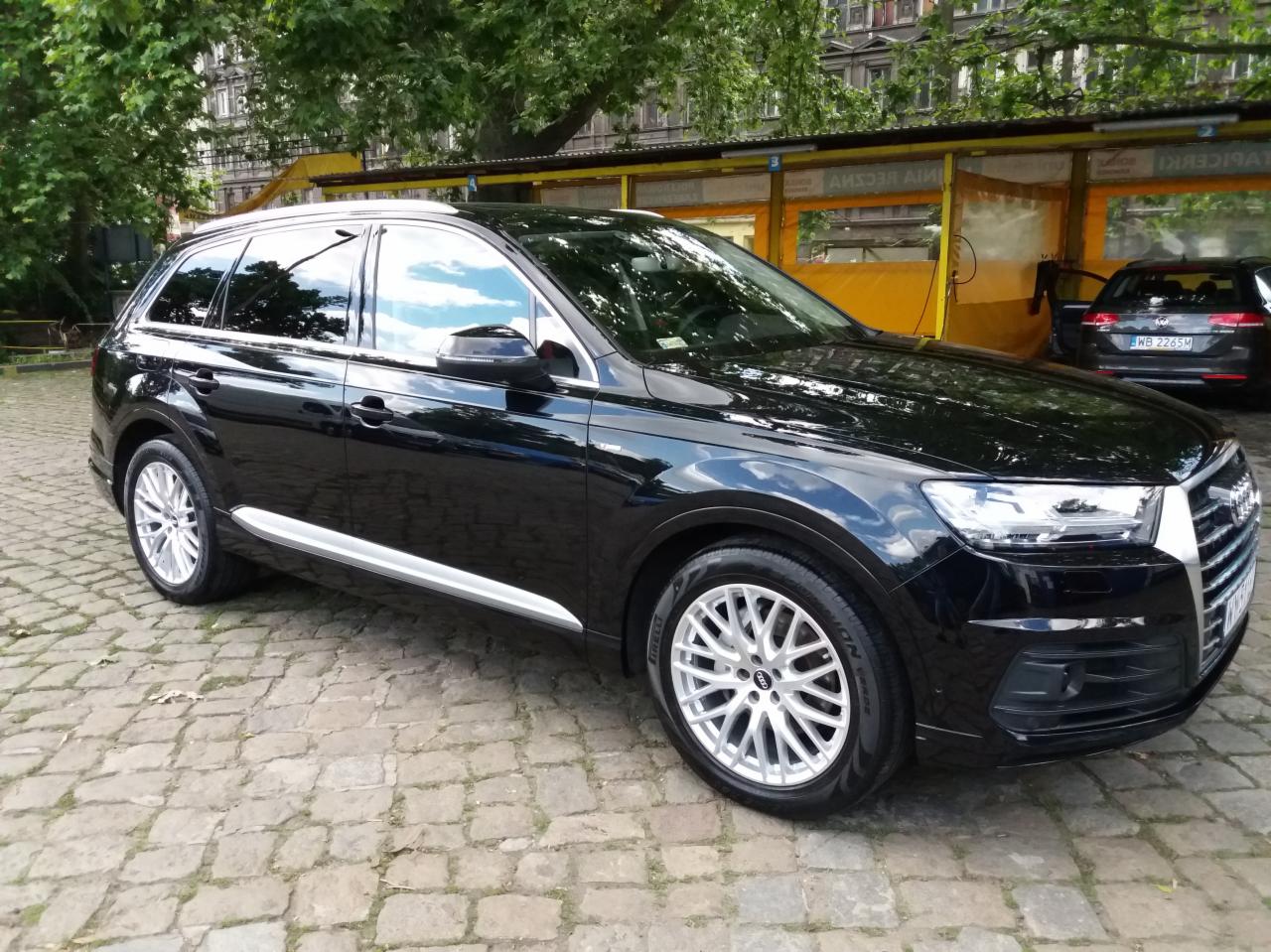 Błyszczący czarny Audi Q7 po umyciu, zaparkowany na brukowanej nawierzchni przed myjnią samochodową z żółtymi elementami.