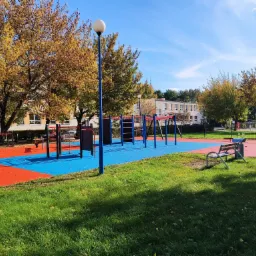 Kolorowy plac zabaw z drabinkami i huśtawkami w Mysłowicach, z bezpieczną nawierzchnią w odcieniach czerwieni i błękitu, oświetlony lampą parkową, otoczony zielonym trawnikiem i drzewami...