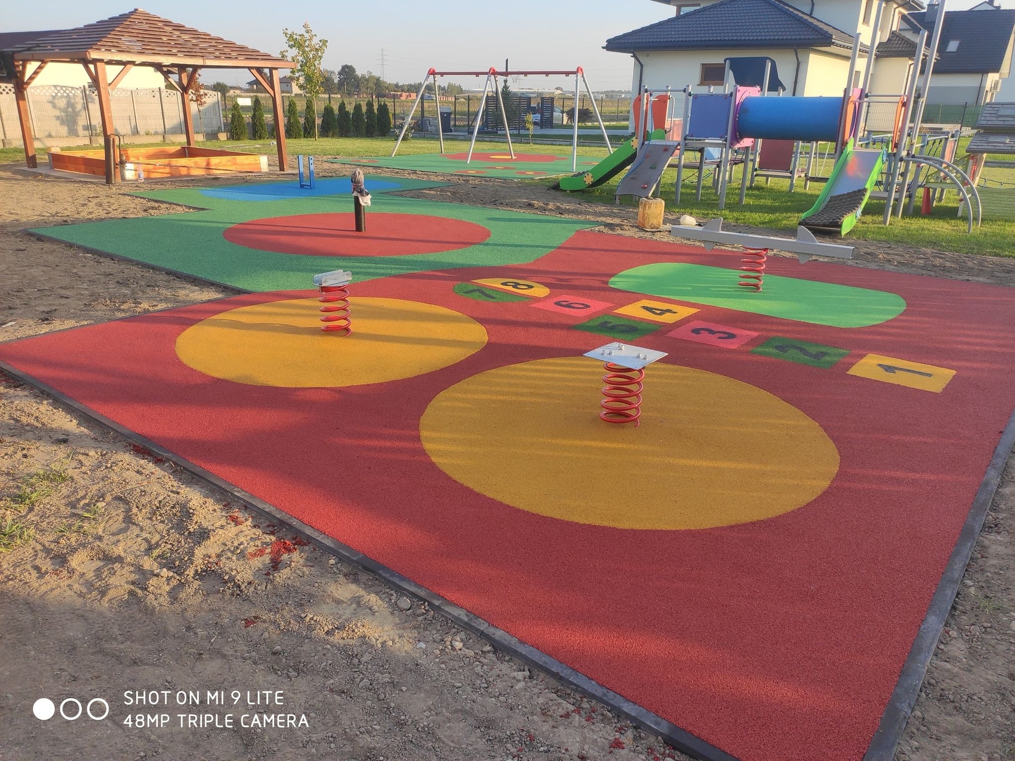 Nowoczesny plac zabaw z kolorową nawierzchnią z gumy, huśtawkami sprężynowymi, drewnianą piaskownicą z zadaszeniem i zestawem zjeżdżalni na tle domów jednorodzinnych.