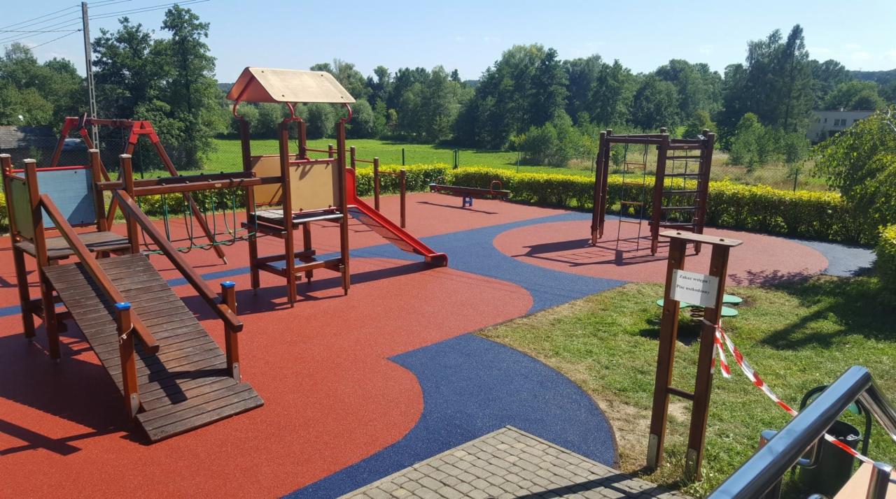 Kompleksowy plac zabaw z czerwoną i niebieską nawierzchnią, zjeżdżalnią, drabinkami i huśtawkami na tle zieleni.