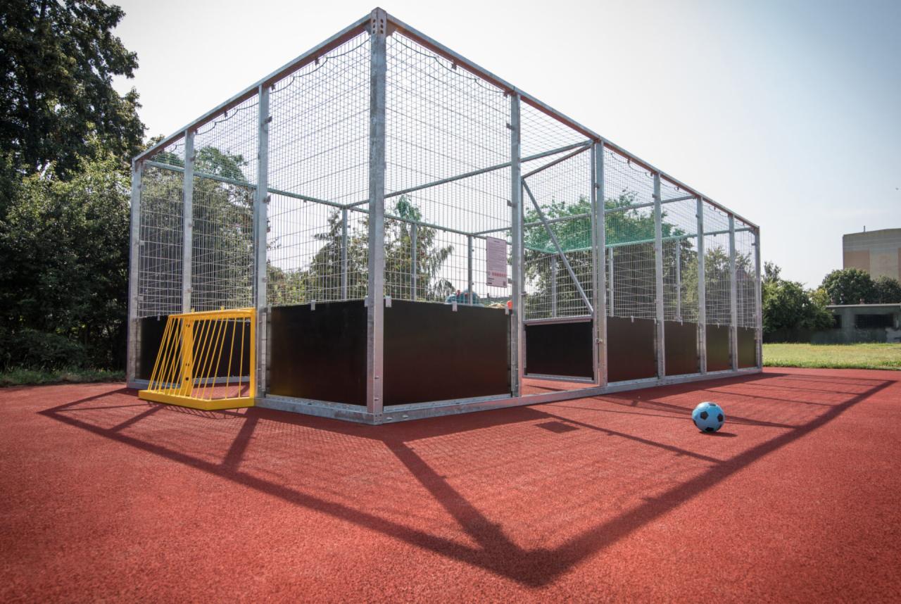 Ogrodzony teren do gry w piłkę nożną na czerwonej, gumowej nawierzchni, z żółtą bramką i niebieską piłką.
