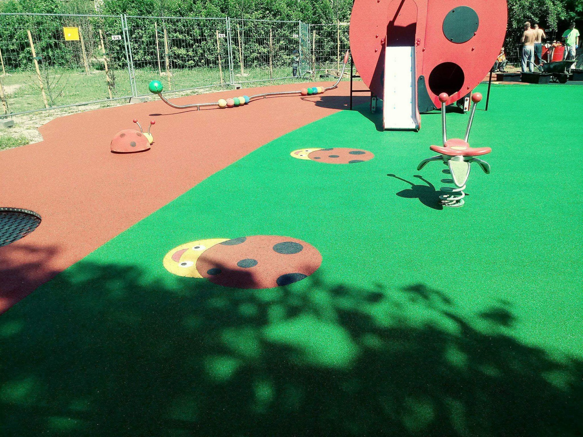 Nowoczesny plac zabaw z zieloną i czerwoną nawierzchnią, wyposażony w dużą czerwoną biedronkę ze zjeżdżalnią, sprężynowca i elementy edukacyjne z koralikami, całość otoczona siatką ogrodzeniową.