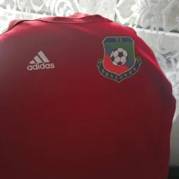 Czerwona koszulka sportowa z logo Adidas i herbem drużyny 'TS SKALANKA' na tle białej firanki