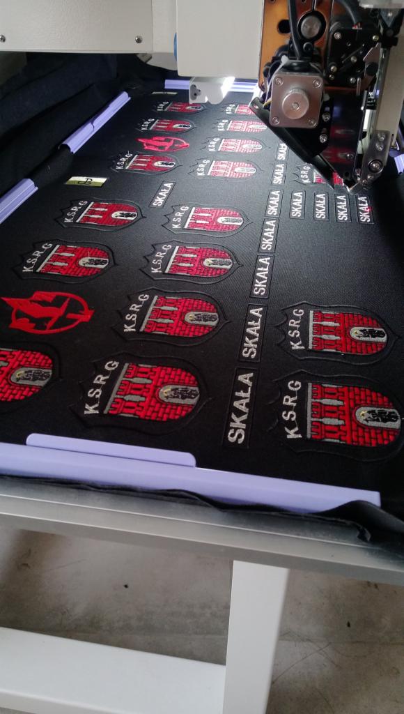 Automatyczny proces tworzenia naszywek z logo i napisem 'SKALA' na czarnym materiale za pomocą maszyny do szycia, widoczne detale igły i mechanizmu maszyny.
