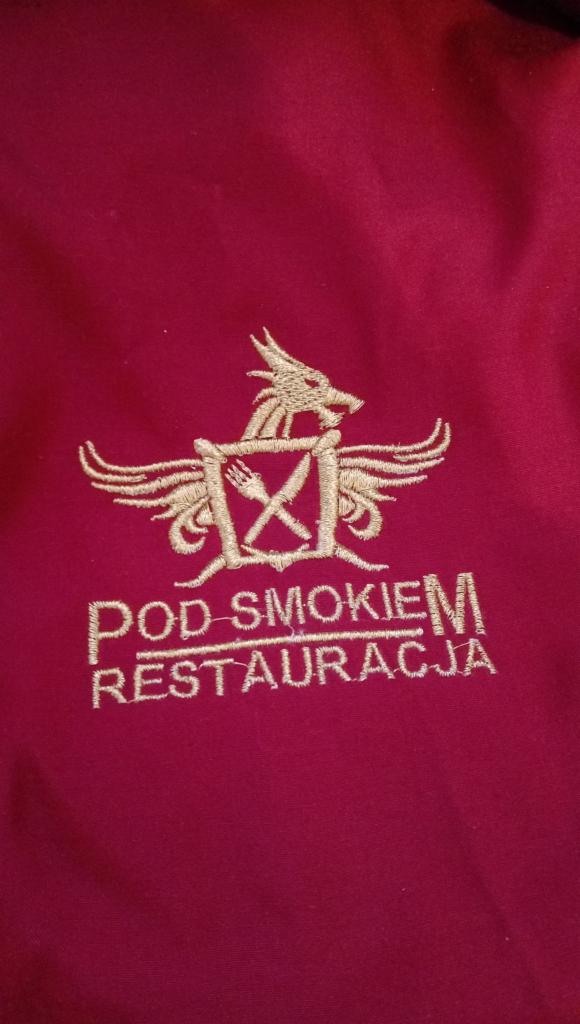 Burgundowa tkanina z wyhaftowanym złotą nicią logo restauracji 'Pod Smokiem' przedstawiającym smoka nad tarczą z skrzyżowanymi sztućcami.