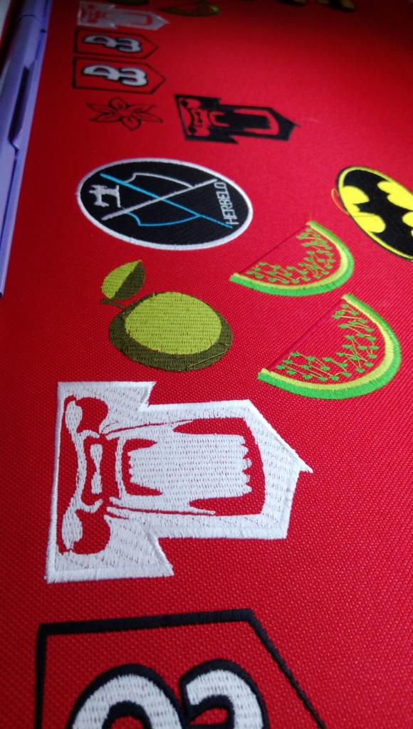 Czerwona tkanina z różnorodnymi, kolorowymi, wyhaftowanymi wzorami, w tym logo, postaci z kreskówek i motywy owocowe.