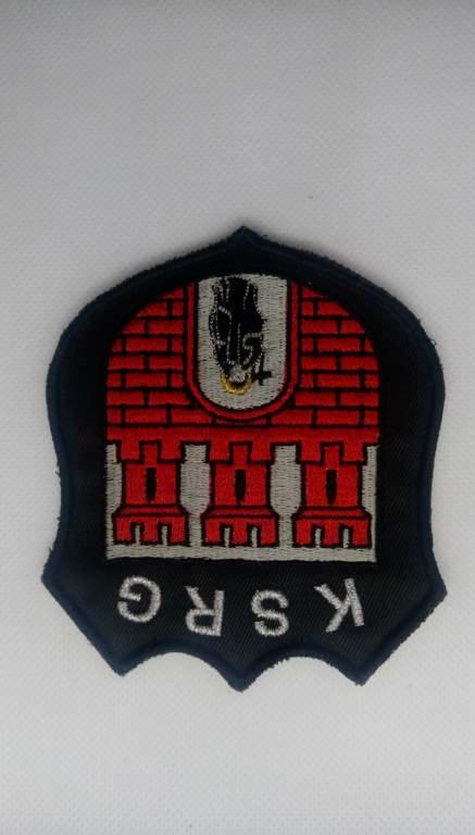 Czarna naszywka z haftowanym herbem przedstawiającym czerwoną ceglaną budowlę z szarymi kolumnami i czarnym symbolem u góry, z białym napisem 'KSRG' u dołu.