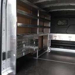 Wnętrze dostawczego samochodu z zabudową warsztatową: metalowe półki z blachy ryflowanej, szuflady z uchwytami i oświetlenie, widok z tyłu przez otwarte drzwi.
