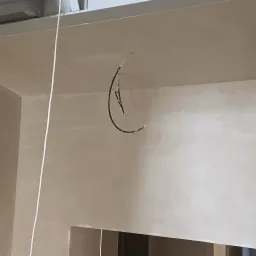 Świeżo nałożony, jasny tynk maszynowy na ścianach i suficie pomieszczenia w stanie surowym; widoczne wystające przewody elektryczne i pionowa biała linka.